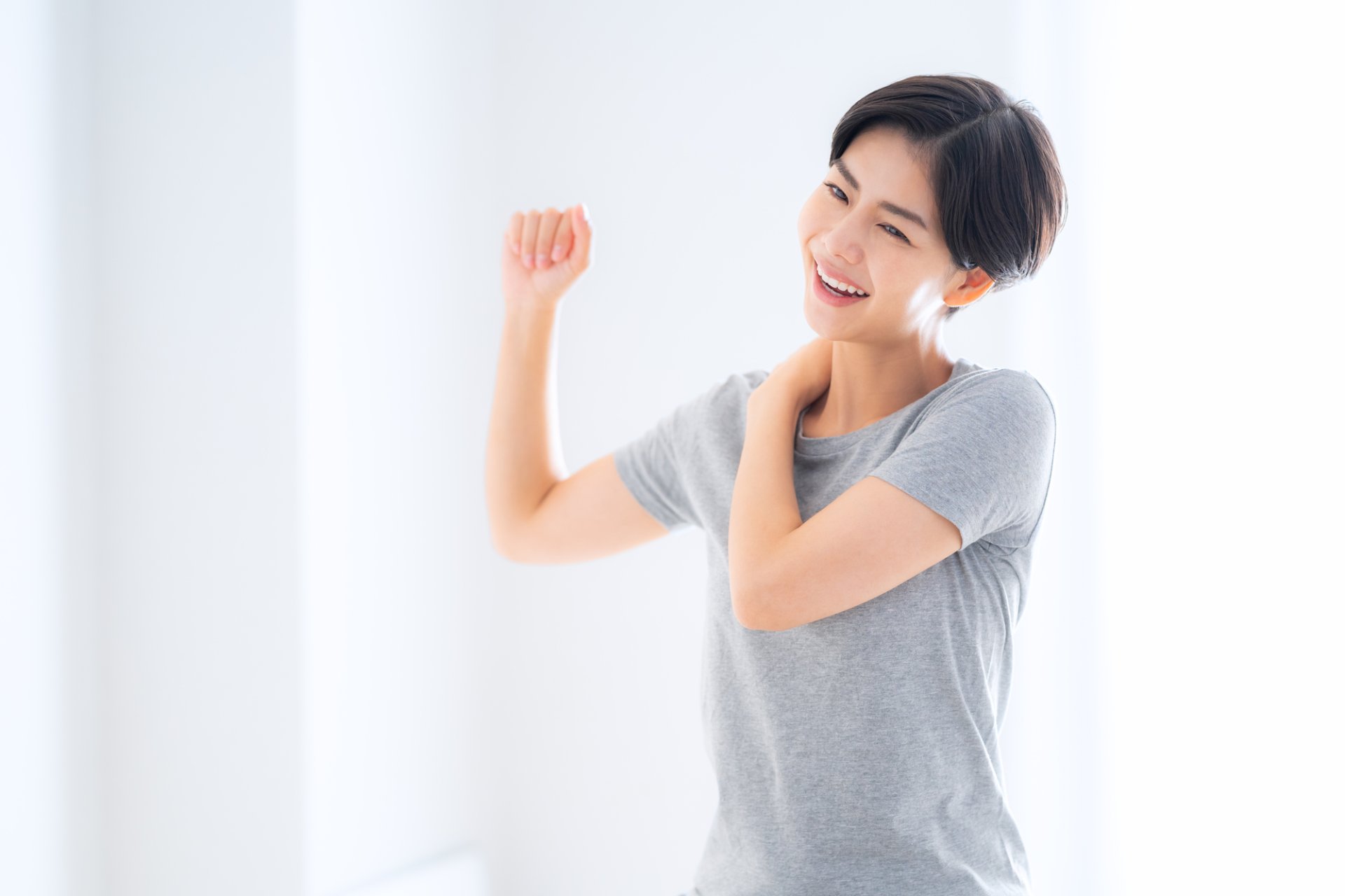 明るい室内で肩に手を当て、笑顔で腕を曲げる短い髪の女性