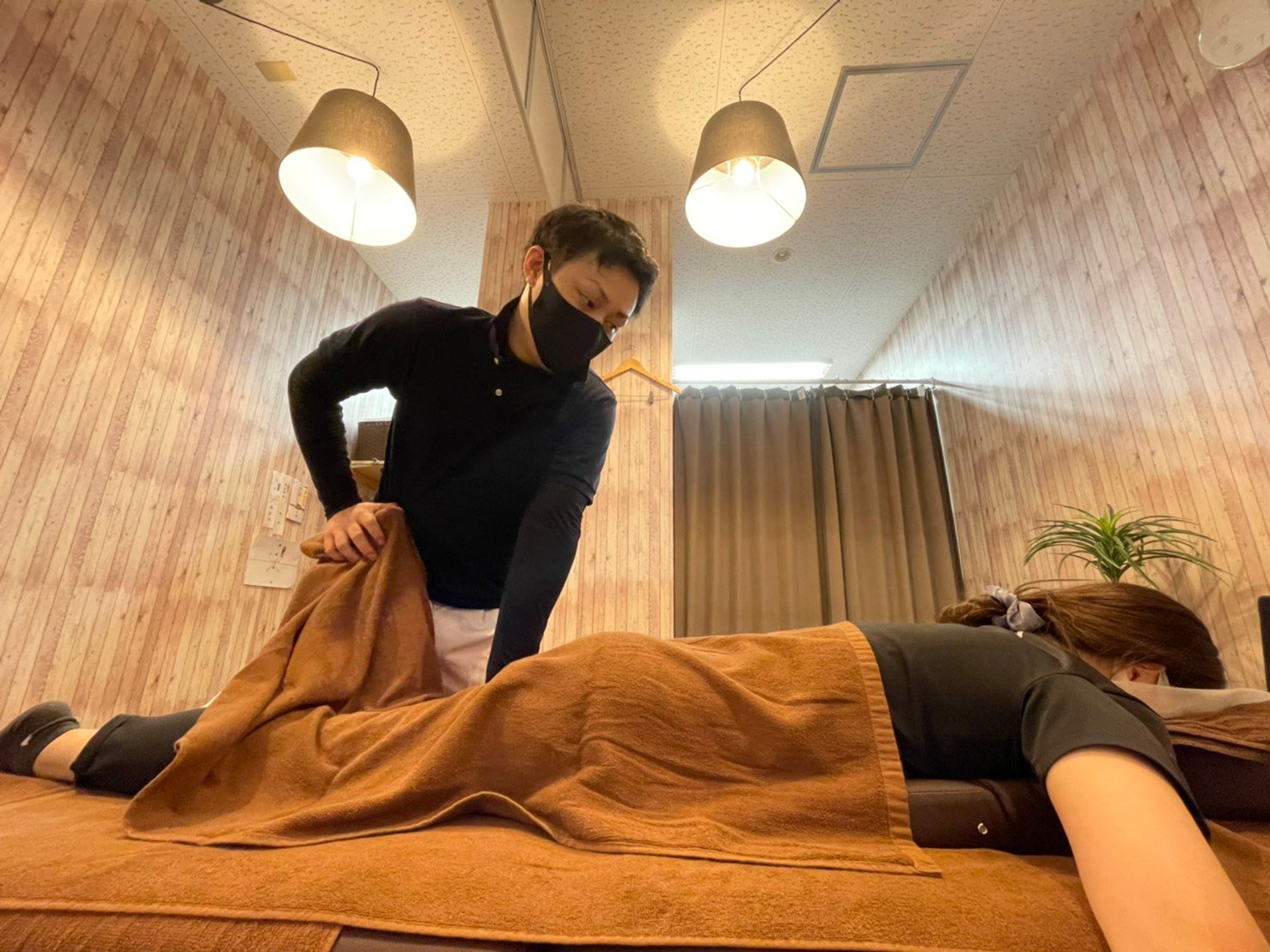木目調の壁とペンダントライトのある施術室で、黒いマスクの施術者がうつ伏せの女性の背中に掛けたタオルを整える様子